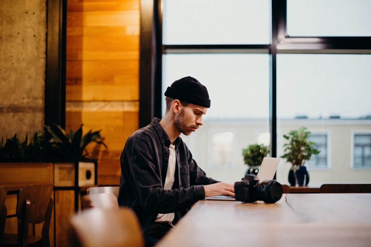 a man sits at a table working on a laptop with a camera beside him