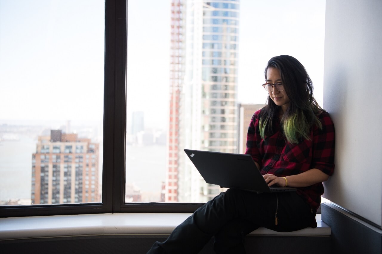 a woman with green hair works on her laptop by a window