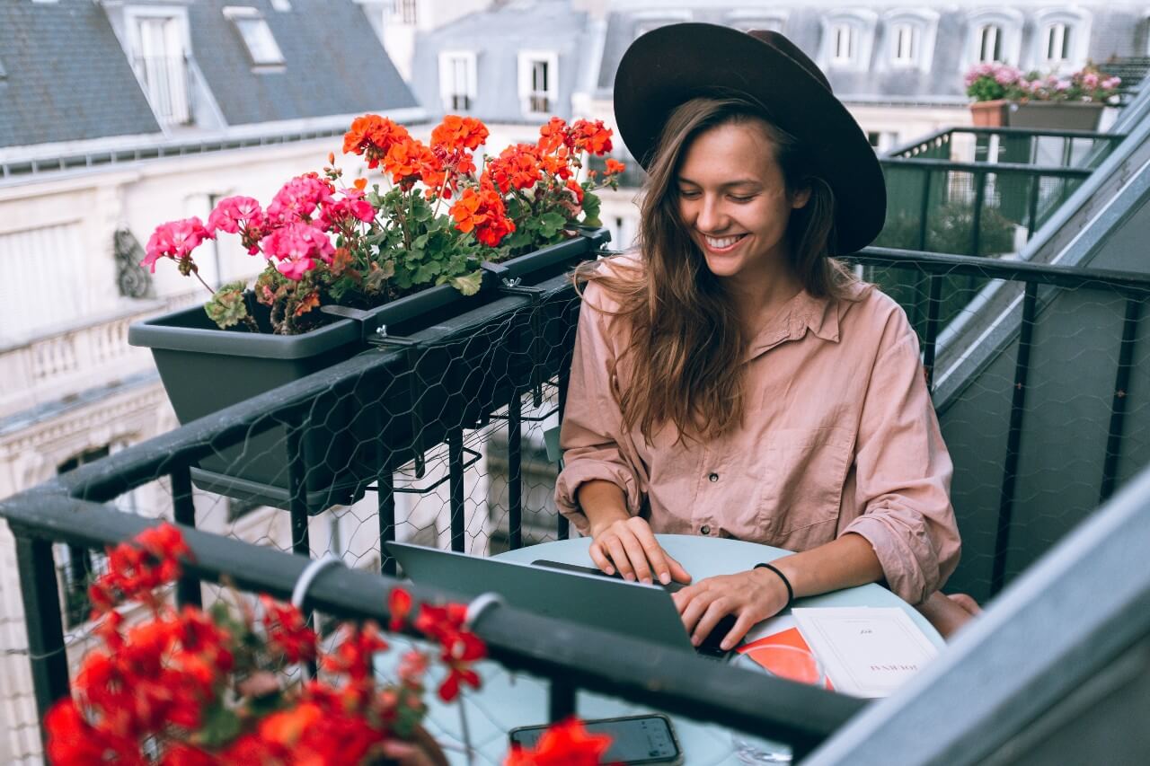 A lady in a pink shirt wearing a hat works at her laptop outside on a balcony