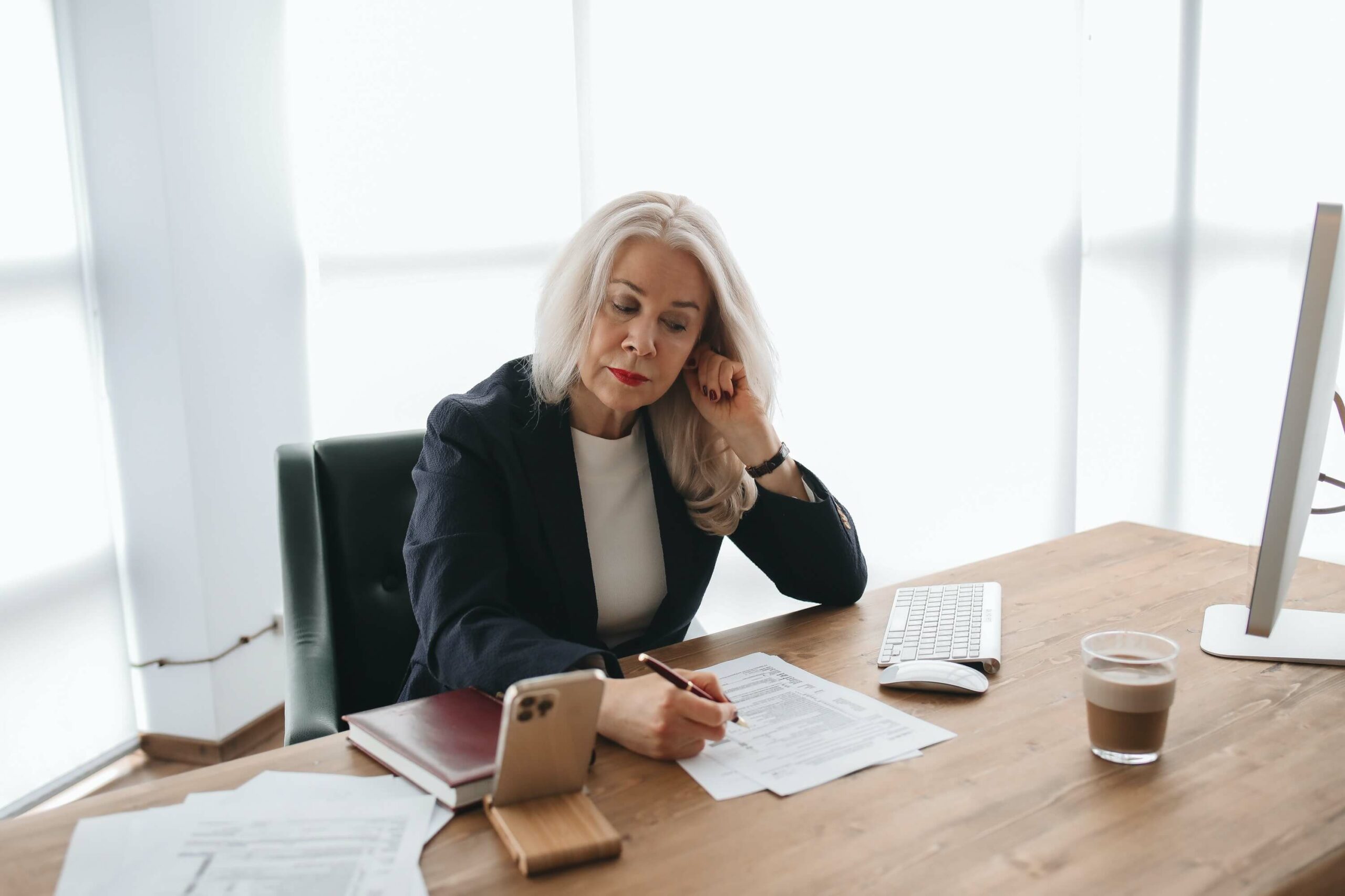 A lady with white hair wearing a black blazer works on paperwork at a desk