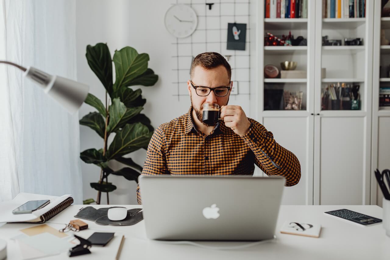 A man in a brown shirt drinks coffee while working on a Macbook