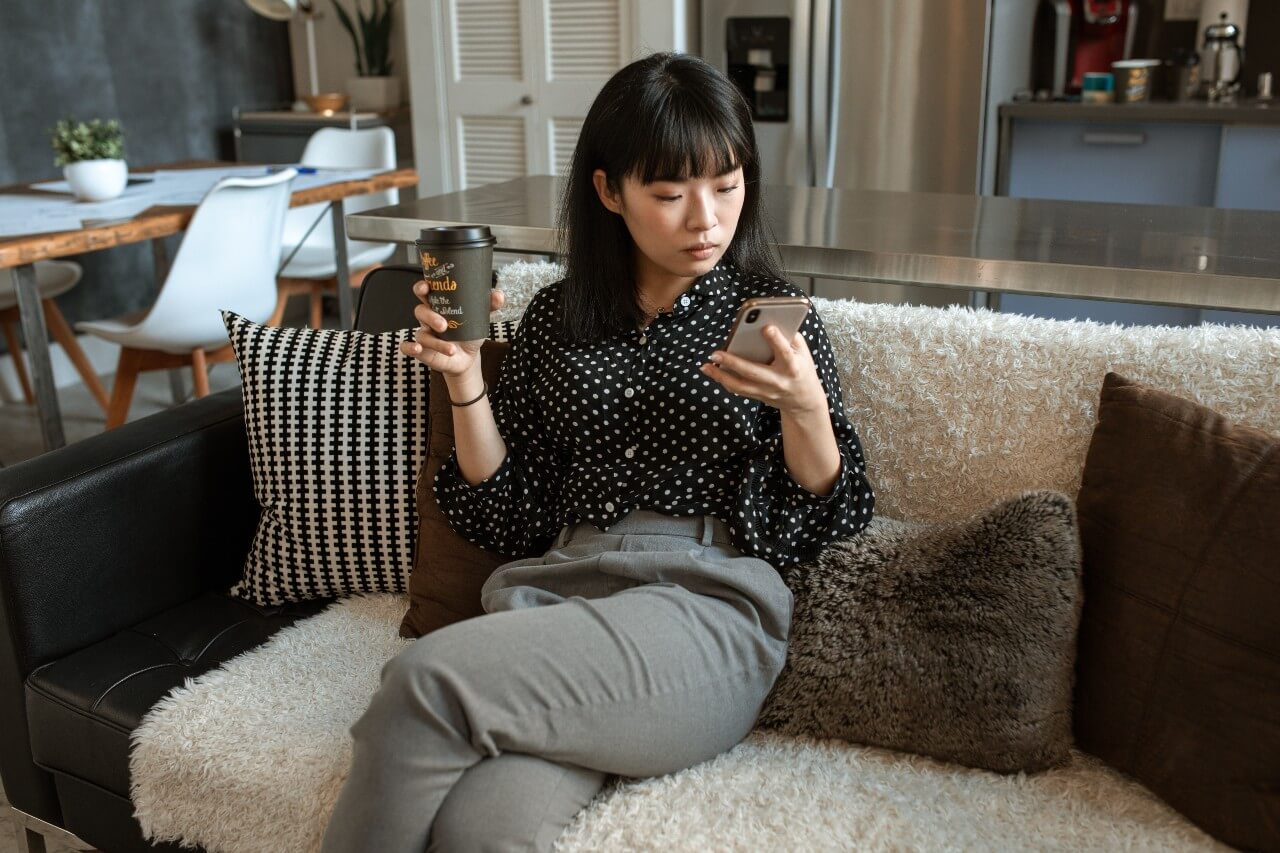 Woman sits on sofa looking at her phone and holding a coffee