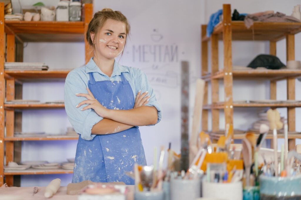 A lady in blue overalls stands in her art studio behind a desk with paintbrushes in pots. She has paint on her face and her blonde hair is tied up.