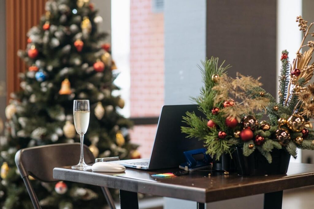 A laptop sits on a desk in front of a Christmas tree, with a glass of sparkling white wine beside it, ready to host a virtual Christmas party