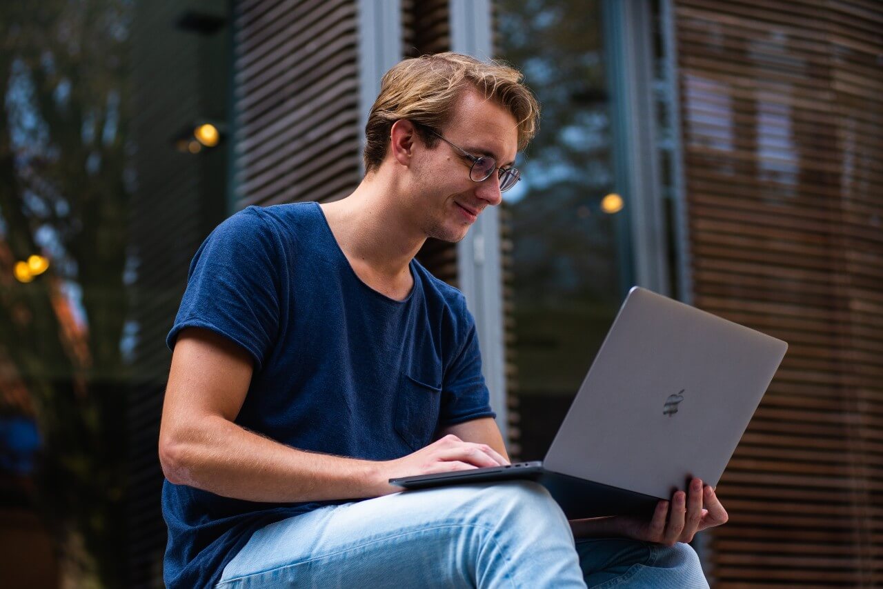 Man wearing blue t shirt and jeans sits with Macbook on his lap, running a credit check on a company