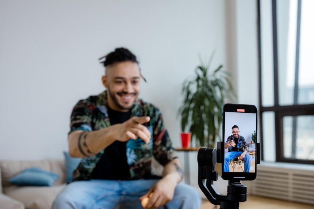 Man with hair tied back wearing a colourful patterned shirt and blue jeans sits on chair in front of mobile phone mounted on a tripod. He is pointing at the camera, while recording a TikTok for his business.