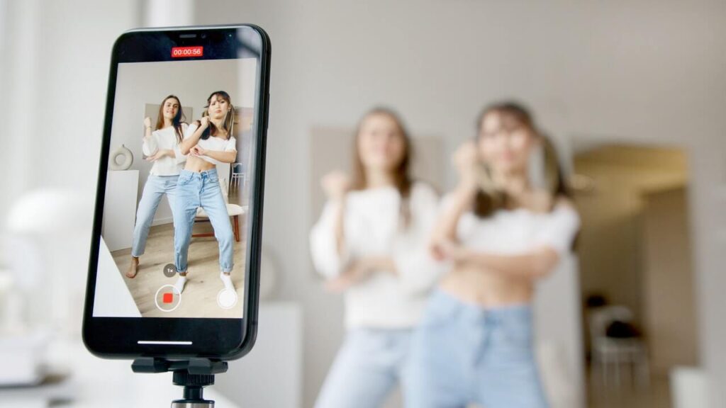 Two women wearing white t shirts and blue jeans stand in front of a mobile phone mounted on a tripod. They are filming a dance as part of a Tiktok marketing video.
